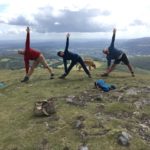 three people doing trichanasana pose on a mountain